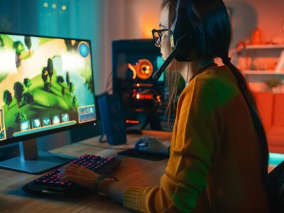 AdobeStock_274386711_Excited and Concentrated Gamer Girl in Glasses and Headset with a Mic Playing Online Strategy Video Game on Her Personal Computer. Room and PC have Colorful Warm Neon Led Lights. Cozy Evening at Home.
